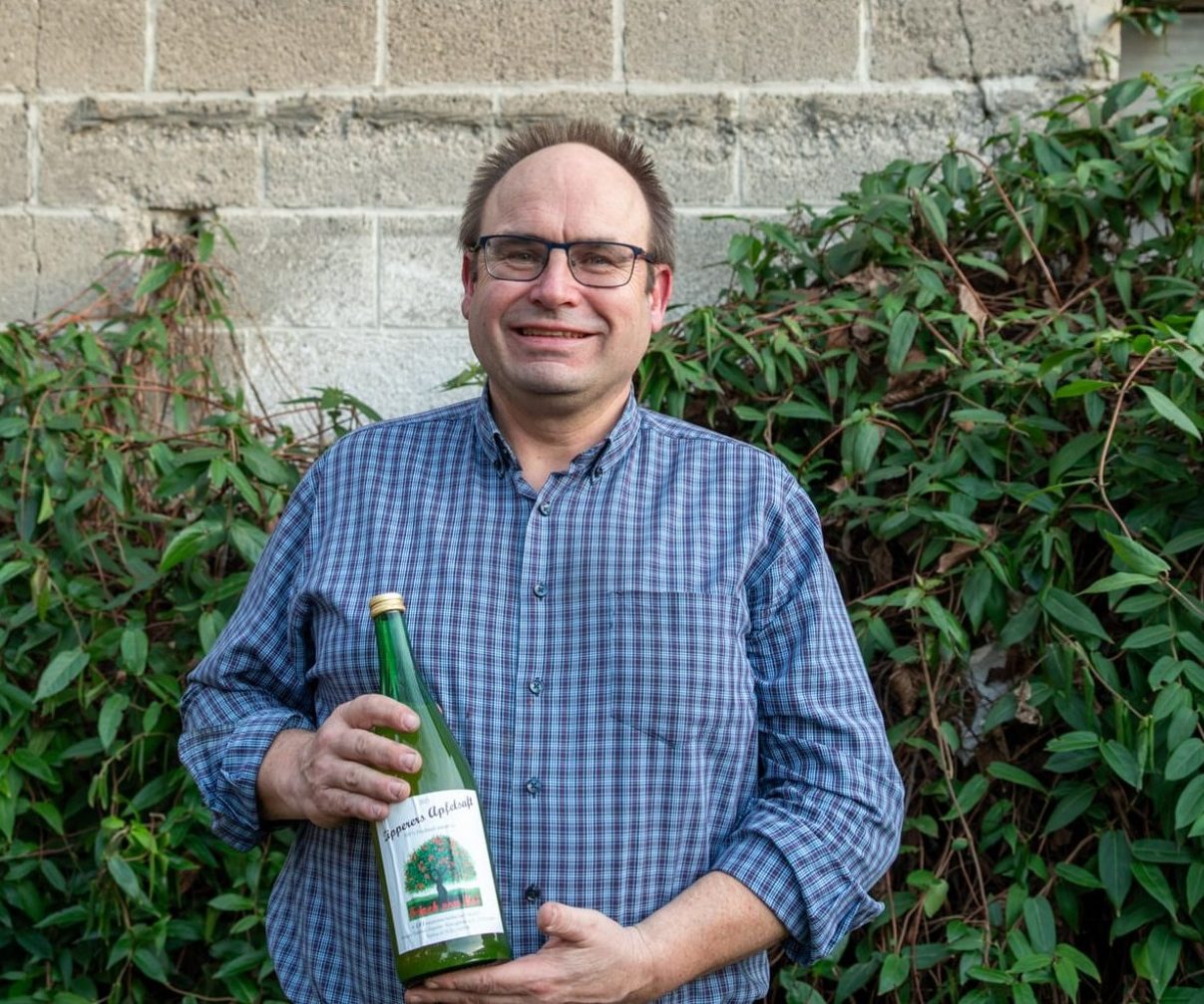 Mann mit Brille hält eine Flasche vor einer grünen Wand aus Pflanzen.