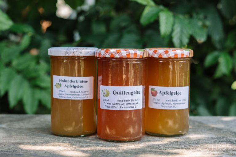 Drei Gläser Marmelade mit Etiketten inmitten von grünen Blättern.